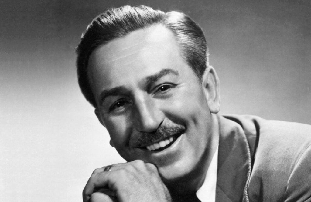 Portrait en noir et blanc d'un homme souriant, moustachu, ressemblant à un personnage de Walt Disney, vêtu d'une veste de costume et posant son menton sur sa main.