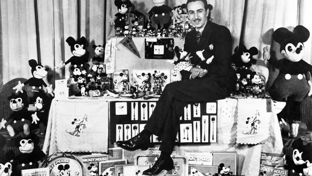 Un homme assis au milieu d'une gamme de produits Mickey Mouse, notamment des jouets en peluche, des figurines et des produits en boîte de Walt Disney, dégageant une attitude fière et joyeuse. Photo en noir et blanc.