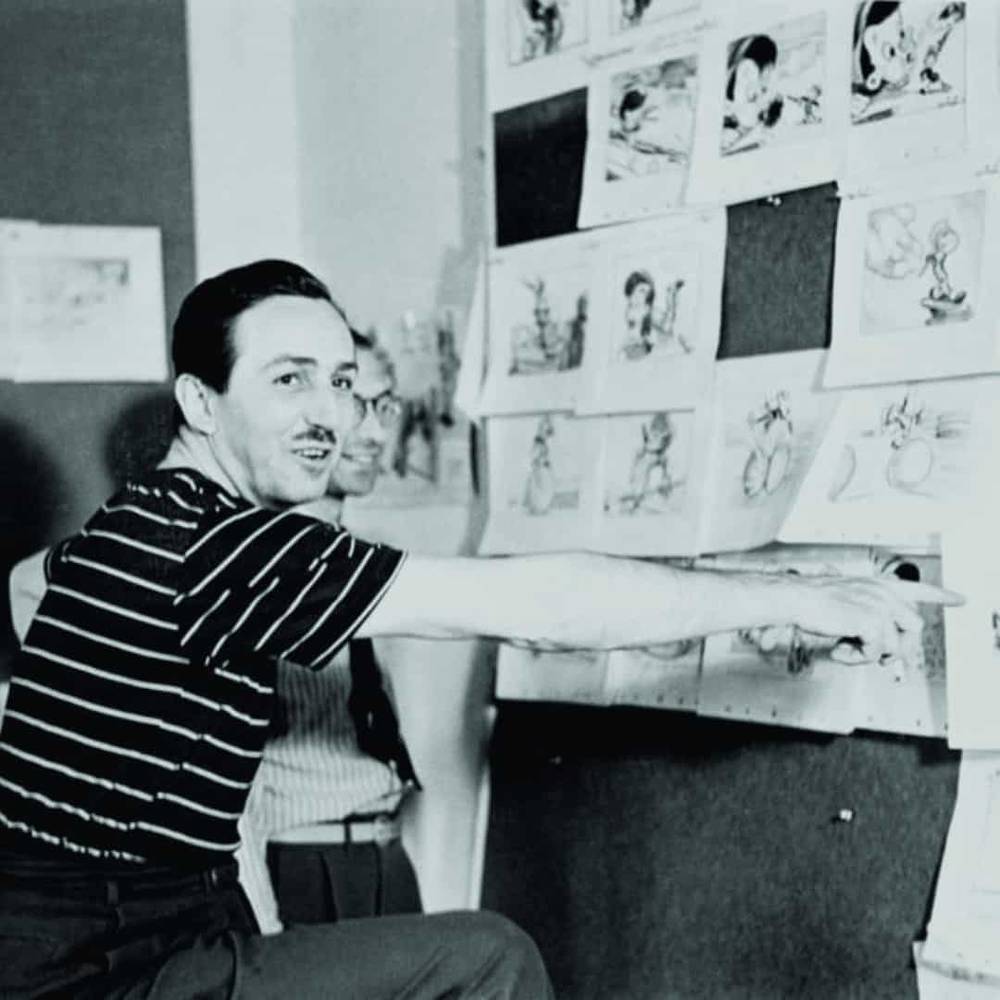 Un homme en chemise rayée montre un storyboard épinglé sur un mur, présentant divers croquis de personnages animés de Walt Disney, exprimant une attitude concentrée et créative.