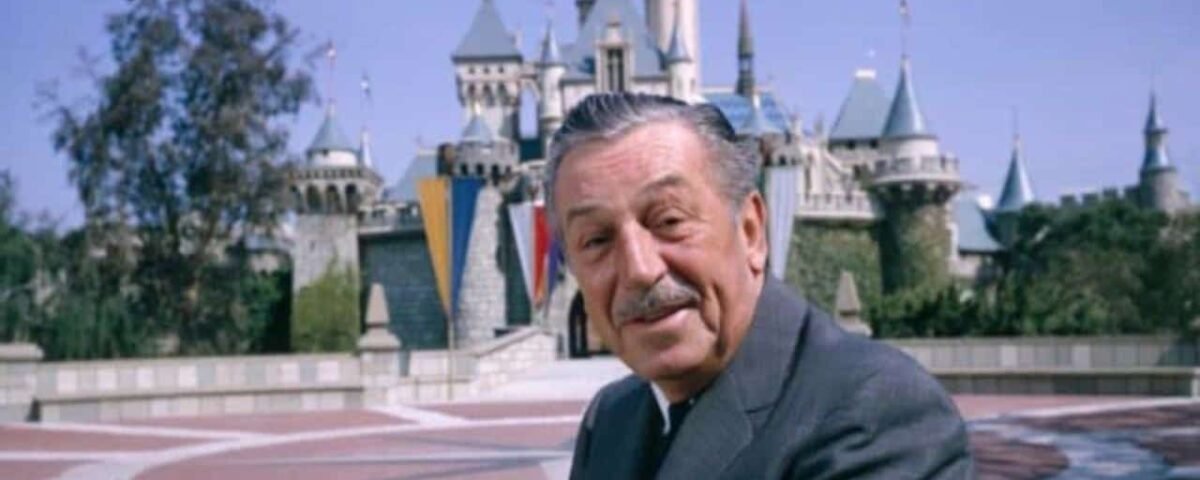 Un homme souriant est assis devant le château de Disneyland, avec un ciel ensoleillé et une verdure vibrante, exprimant une atmosphère accueillante ancrée dans l'industrie du divertissement.