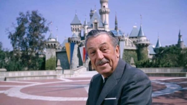 Un homme souriant est assis devant le château de Disneyland, avec un ciel ensoleillé et une verdure vibrante, exprimant une atmosphère accueillante ancrée dans l'industrie du divertissement.