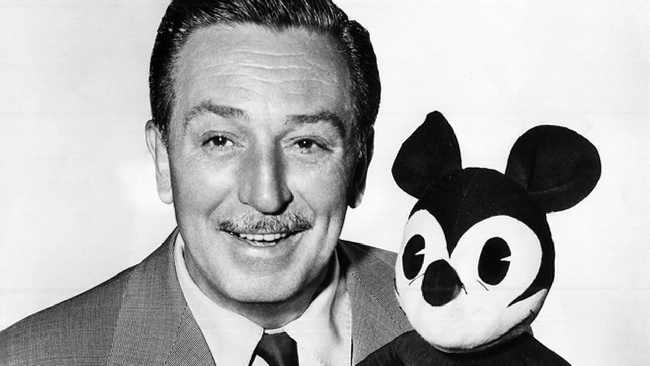 Photo en noir et blanc d’un homme souriant tenant une peluche de personnage de dessin animé, vêtu d’un costume et d’une cravate. La peluche ressemble à un animal stylisé avec de grandes oreilles et de grands yeux utilisé dans un quiz vrai/faux