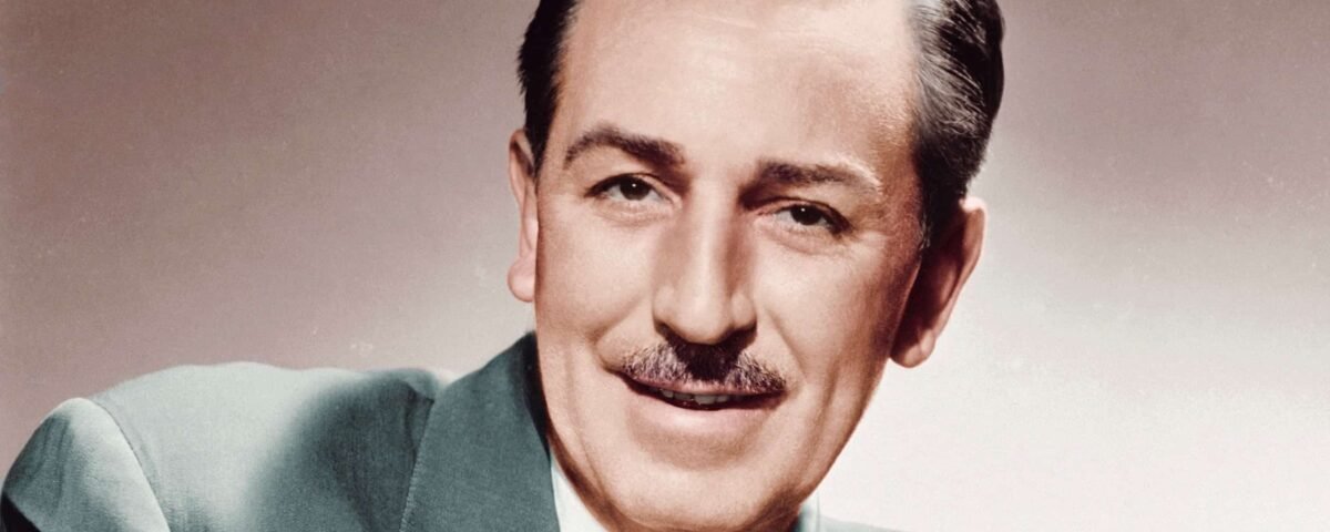 Portrait d'un homme d'âge moyen souriant avec une moustache soignée, vêtu d'un costume vert et d'une cravate à motifs, sur un fond sourd rappelant les studios d'animation de Walt Disney.