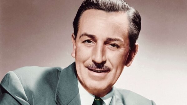 Portrait d'un homme d'âge moyen souriant avec une moustache soignée, vêtu d'un costume vert et d'une cravate à motifs, sur un fond sourd rappelant les studios d'animation de Walt Disney.
