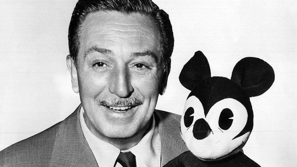 Une photo en noir et blanc d’un homme souriant en costume tenant une peluche souris de style Walt Disney.