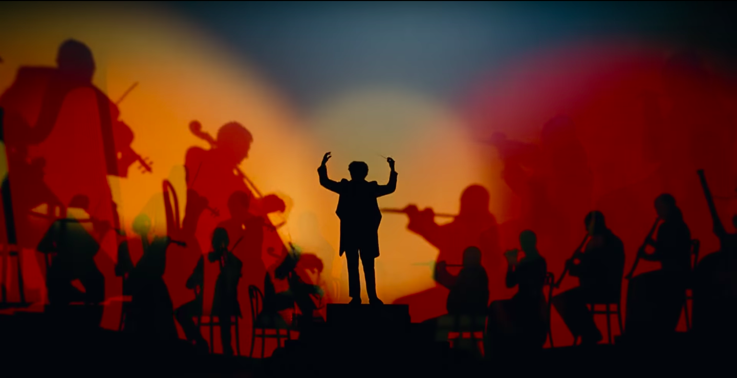 Chef d'orchestre et musiciens silhouettés se produisant sur scène dans une fantaisie de lumières colorées dans les tons rouge et jaune.