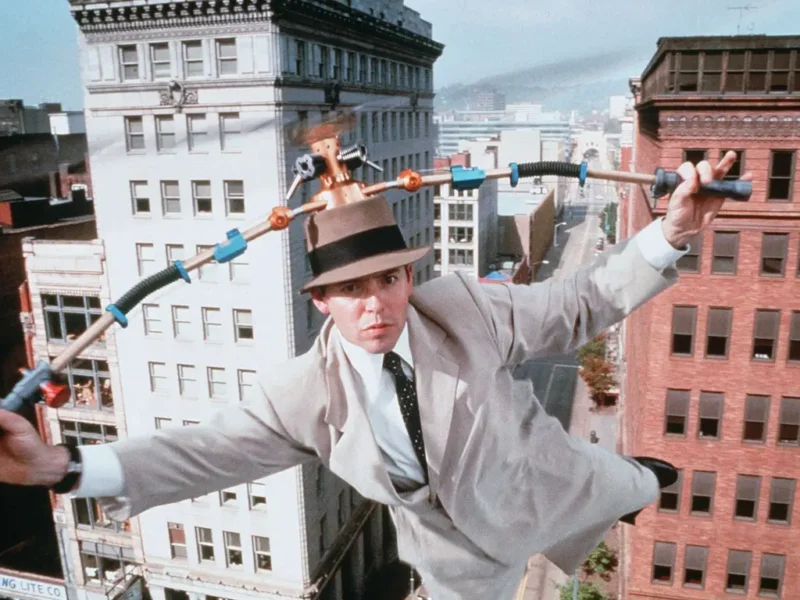 Un homme portant un fedora exécute un numéro d'équilibriste sur un fil électrique entre les bâtiments, utilisant un engin à plusieurs poignées pour maintenir l'équilibre lors de l'événement "Le Noël de Mickey".