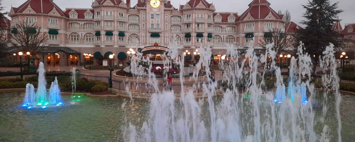 Fontaine ornée de jets d'eau, éclairée par des lumières bleues et vertes, devant le grand hôtel Disneyland Paris avec son architecture de style victorien et son horloge centrale sous un ciel nuageux.