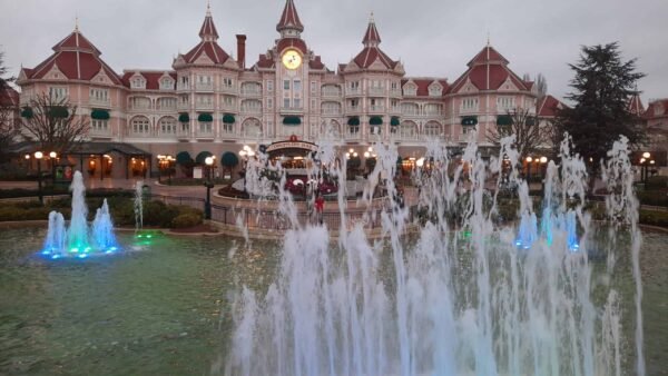 Fontaine ornée de jets d'eau, éclairée par des lumières bleues et vertes, devant le grand hôtel Disneyland Paris avec son architecture de style victorien et son horloge centrale sous un ciel nuageux.