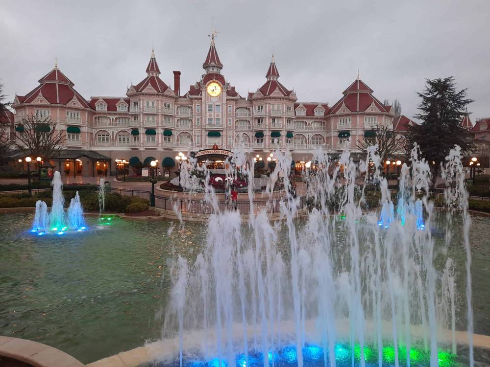 Fontaine ornée de jets d'eau, éclairée par des lumières bleues et vertes, devant le grand hôtel Disneyland Paris avec son architecture de style victorien et son horloge centrale sous un ciel nuageux.