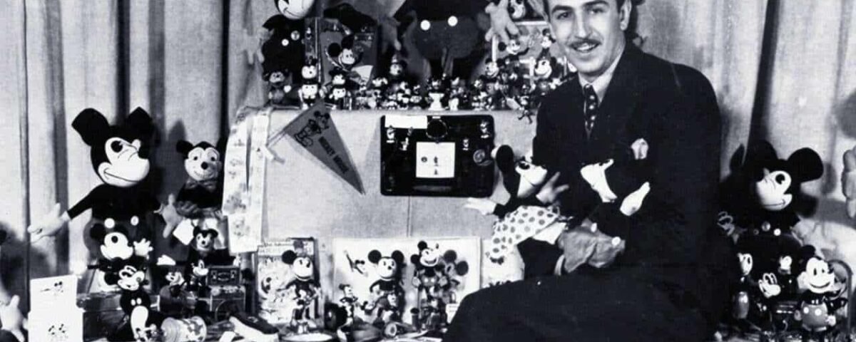 Une photo en noir et blanc d'un homme entouré d'une grande collection de jouets Mickey Mouse et d'articles Walt Disney, affichant divers objets avec un sourire fier.