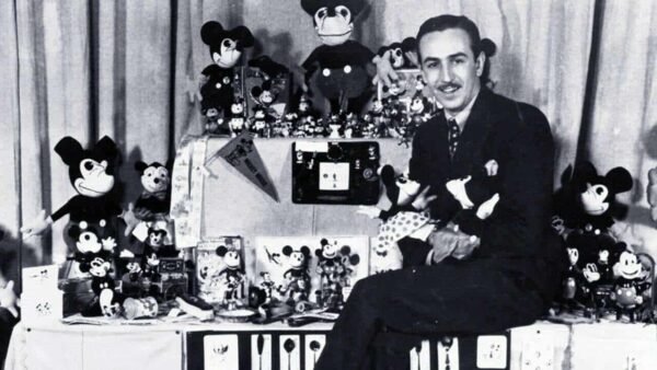 Une photo en noir et blanc d'un homme entouré d'une grande collection de jouets Mickey Mouse et d'articles Walt Disney, affichant divers objets avec un sourire fier.