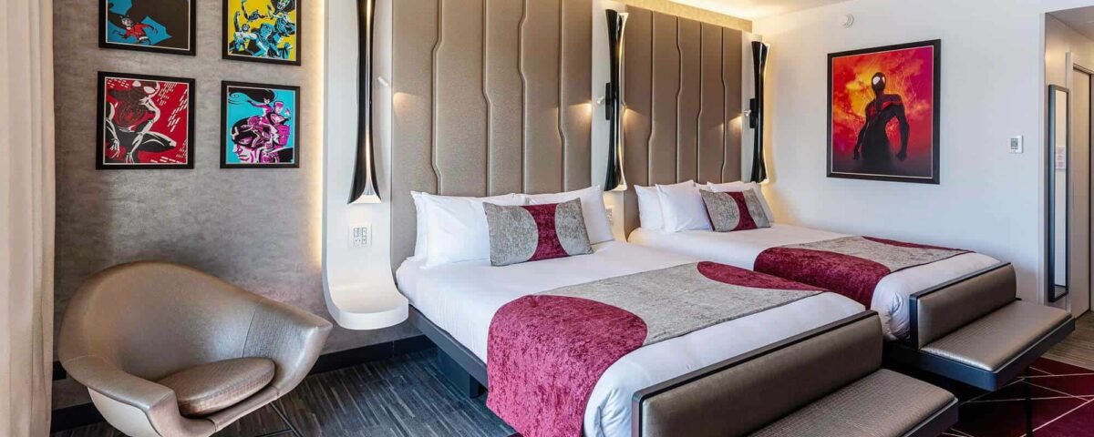 Une chambre d'hôtel moderne près de Disneyland Paris avec deux lits, une literie marron et grise, des œuvres d'art vibrantes sur les murs, une grande chaise grise courbée et un tapis à motifs géométriques.