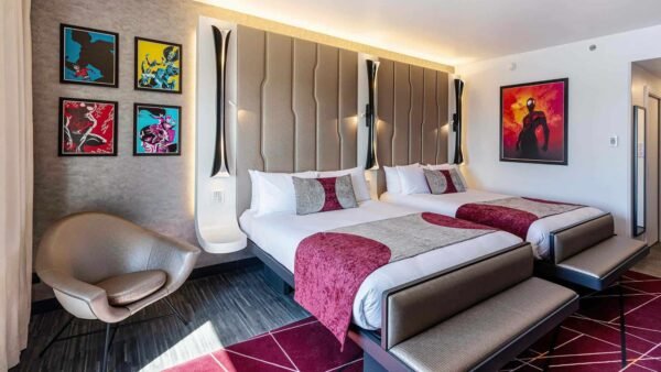 Une chambre d'hôtel moderne près de Disneyland Paris avec deux lits, une literie marron et grise, des œuvres d'art vibrantes sur les murs, une grande chaise grise courbée et un tapis à motifs géométriques.