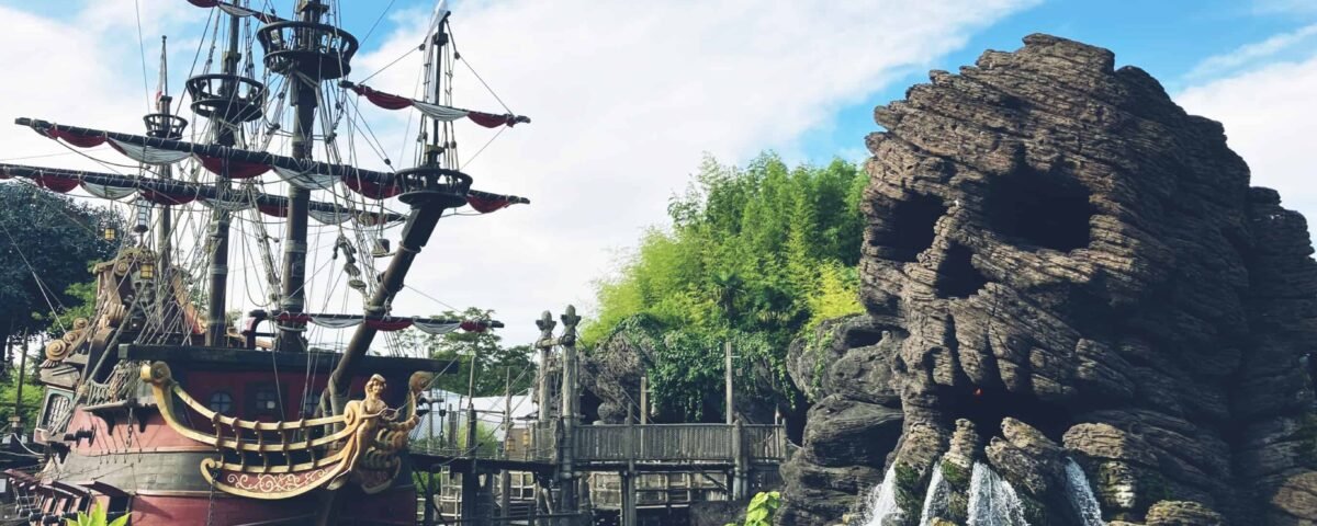 Une zone imaginative sur le thème des pirates à Disneyland avec un grand bateau pirate détaillé ancré à côté d'une formation rocheuse accidentée en forme de crâne avec des cascades. Une passerelle en bois et de la verdure rehaussent la vue panoramique.