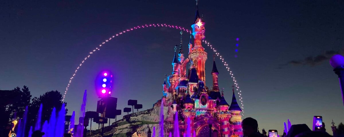Vue nocturne d'un château bien éclairé à Disneyland avec une foule de personnes âgées profitant du spectacle de lumière. Un bel arc de lumière balaye le château, créant une atmosphère magique.