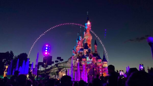 Vue nocturne d'un château bien éclairé à Disneyland avec une foule de personnes âgées profitant du spectacle de lumière. Un bel arc de lumière balaye le château, créant une atmosphère magique.