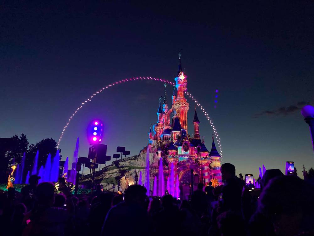 Vue nocturne d'un château bien éclairé à Disneyland avec une foule de personnes âgées profitant du spectacle de lumière. Un bel arc de lumière balaye le château, créant une atmosphère magique.