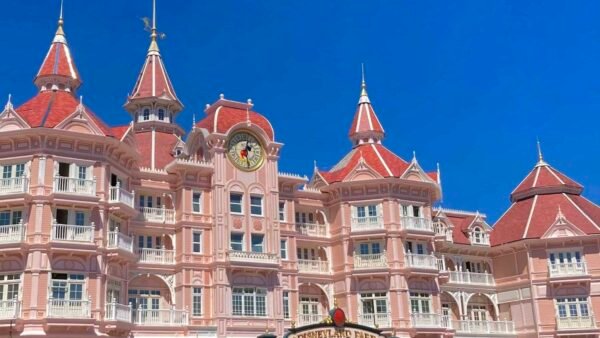 Une image vibrante de l'Hôtel Disney, mettant en valeur la façade rose et les toits ornés d'un bâtiment inspiré des contes de fées sous un ciel bleu clair.