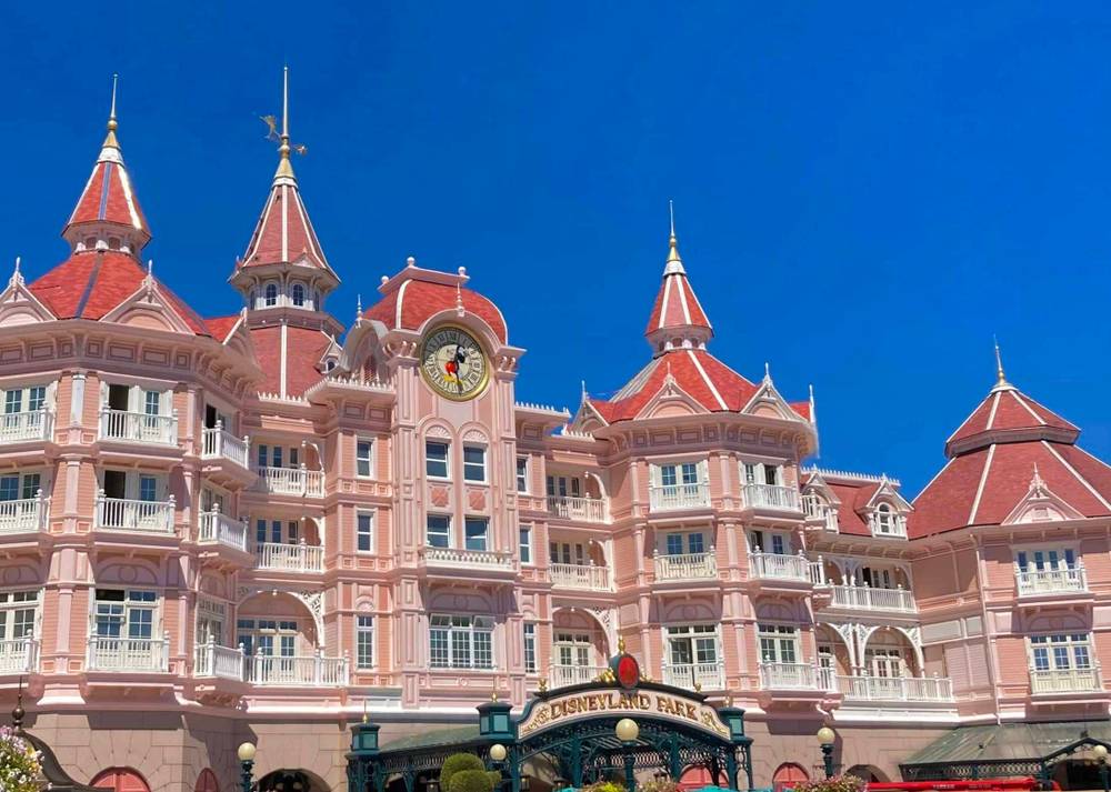 Une image vibrante de l'Hôtel Disney, mettant en valeur la façade rose et les toits ornés d'un bâtiment inspiré des contes de fées sous un ciel bleu clair.