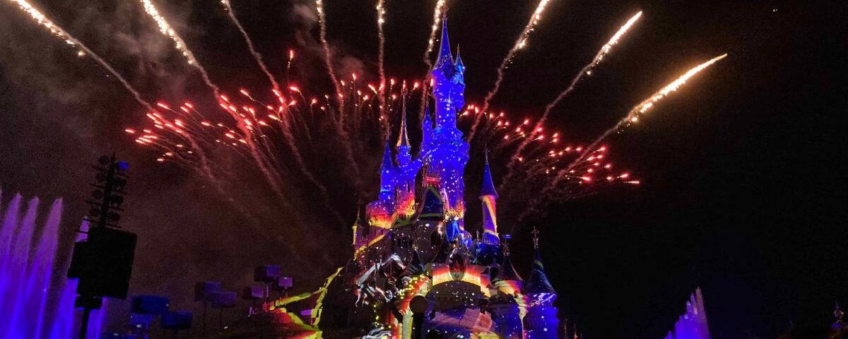 Un spectacle nocturne à Disneyland Paris mettant en vedette un château bien illuminé avec des feux d'artifice vibrants éclatant dans le ciel sombre au-dessus, tandis qu'une foule de spectateurs regarde avec admiration.