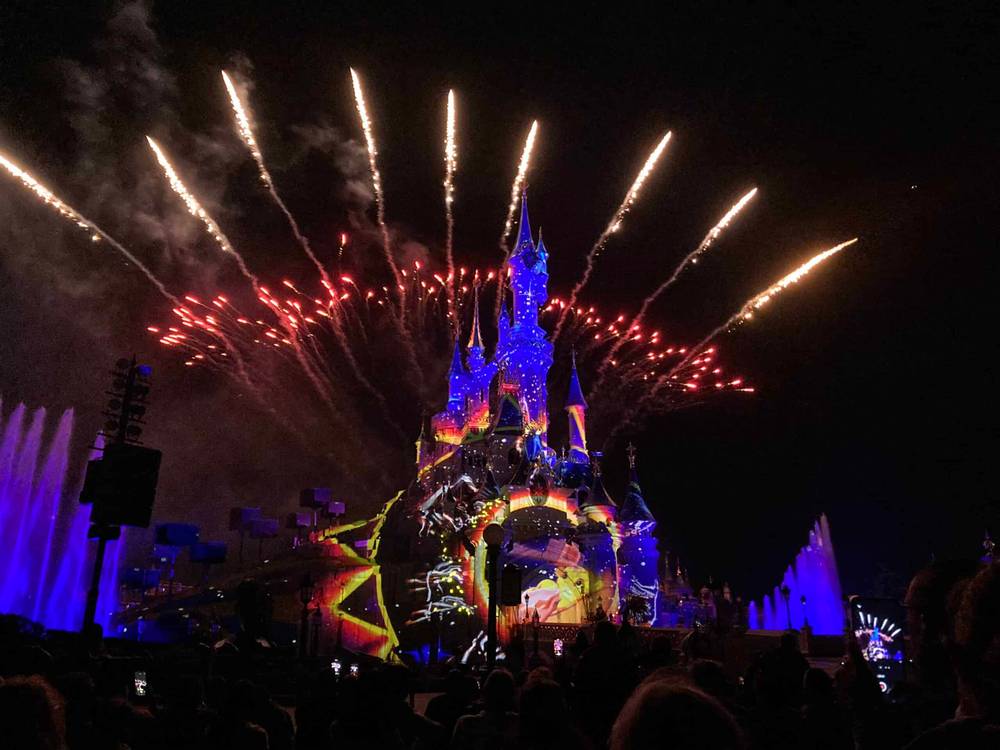 Un spectacle nocturne à Disneyland Paris mettant en vedette un château bien illuminé avec des feux d'artifice vibrants éclatant dans le ciel sombre au-dessus, tandis qu'une foule de spectateurs regarde avec admiration.