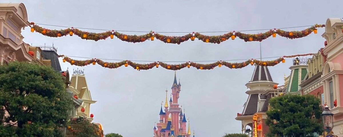 Des décorations d'automne sont suspendues au-dessus d'une rue bordée de bâtiments colorés menant à Disneyland Paris par temps couvert.