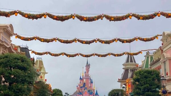 Des décorations d'automne sont suspendues au-dessus d'une rue bordée de bâtiments colorés menant à Disneyland Paris par temps couvert.