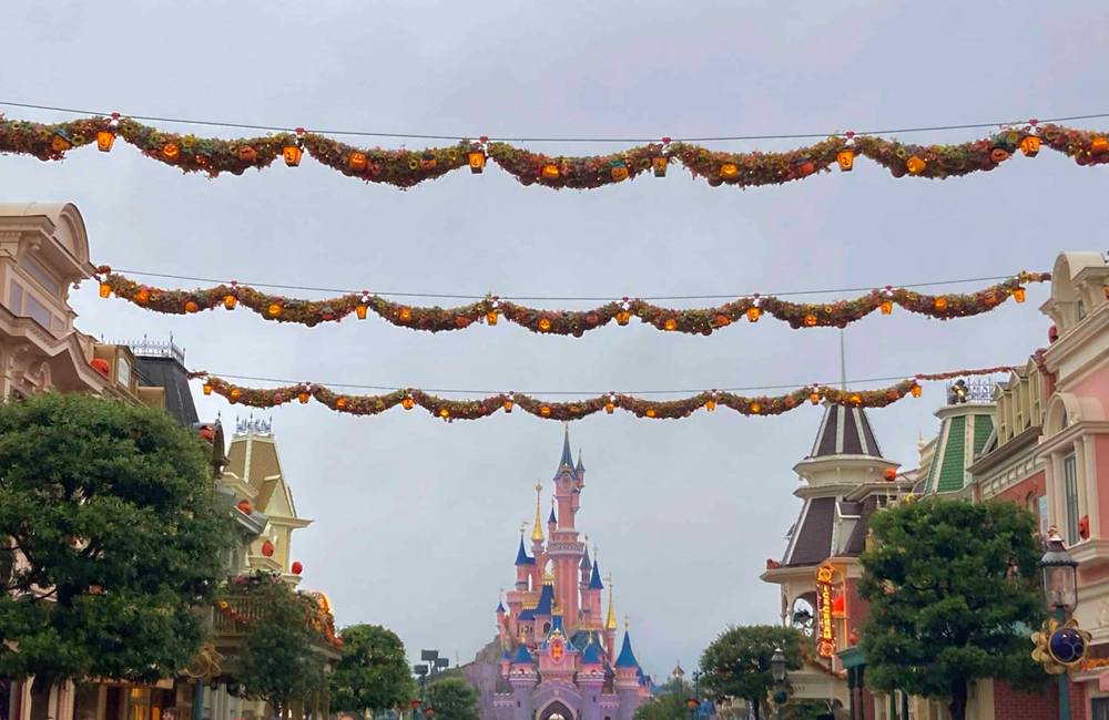 Des décorations d'automne sont suspendues au-dessus d'une rue bordée de bâtiments colorés menant à Disneyland Paris par temps couvert.