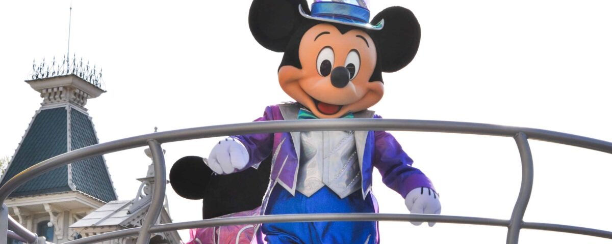 Mickey Mouse, vêtu d'un costume bleu étincelant et d'un haut-de-forme, salue du haut d'un char avec un château en toile de fond à Disneyland Paris dans un décor de défilé.