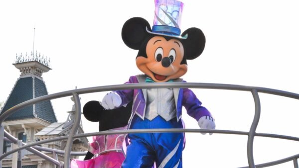 Mickey Mouse, vêtu d'un costume bleu étincelant et d'un haut-de-forme, salue du haut d'un char avec un château en toile de fond à Disneyland Paris dans un décor de défilé.
