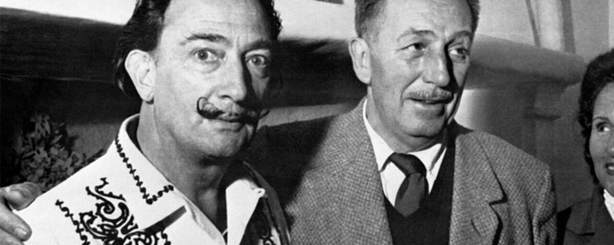 Deux hommes souriants, l'un avec une moustache ornée rappelant Salvador Dali, portant une chemise décorée, et l'autre en costume-cravate, se tenant ensemble lors d'un événement en salle.