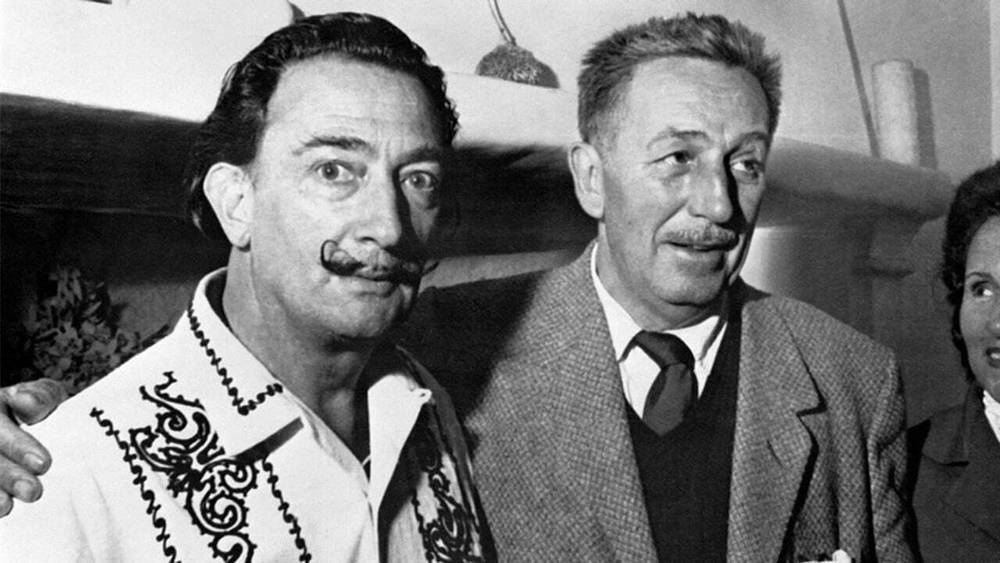 Deux hommes souriants, l'un avec une moustache ornée rappelant Salvador Dali, portant une chemise décorée, et l'autre en costume-cravate, se tenant ensemble lors d'un événement en salle.
