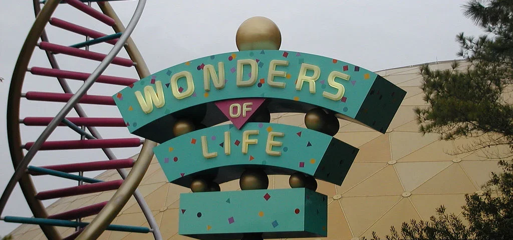 Un panneau coloré indiquant « merveilles de la vie » dans un parc à thème, placé contre un grand dôme doré et entouré de structures ressemblant à des brins d'ADN et d'arbres sous un ciel nuageux.