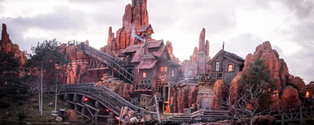 Une vue panoramique d'un paysage montagneux et accidenté avec un village en bois et des ponts au crépuscule, se reflétant dans un lac tranquille en contrebas, rappelant Disneyland Paris.