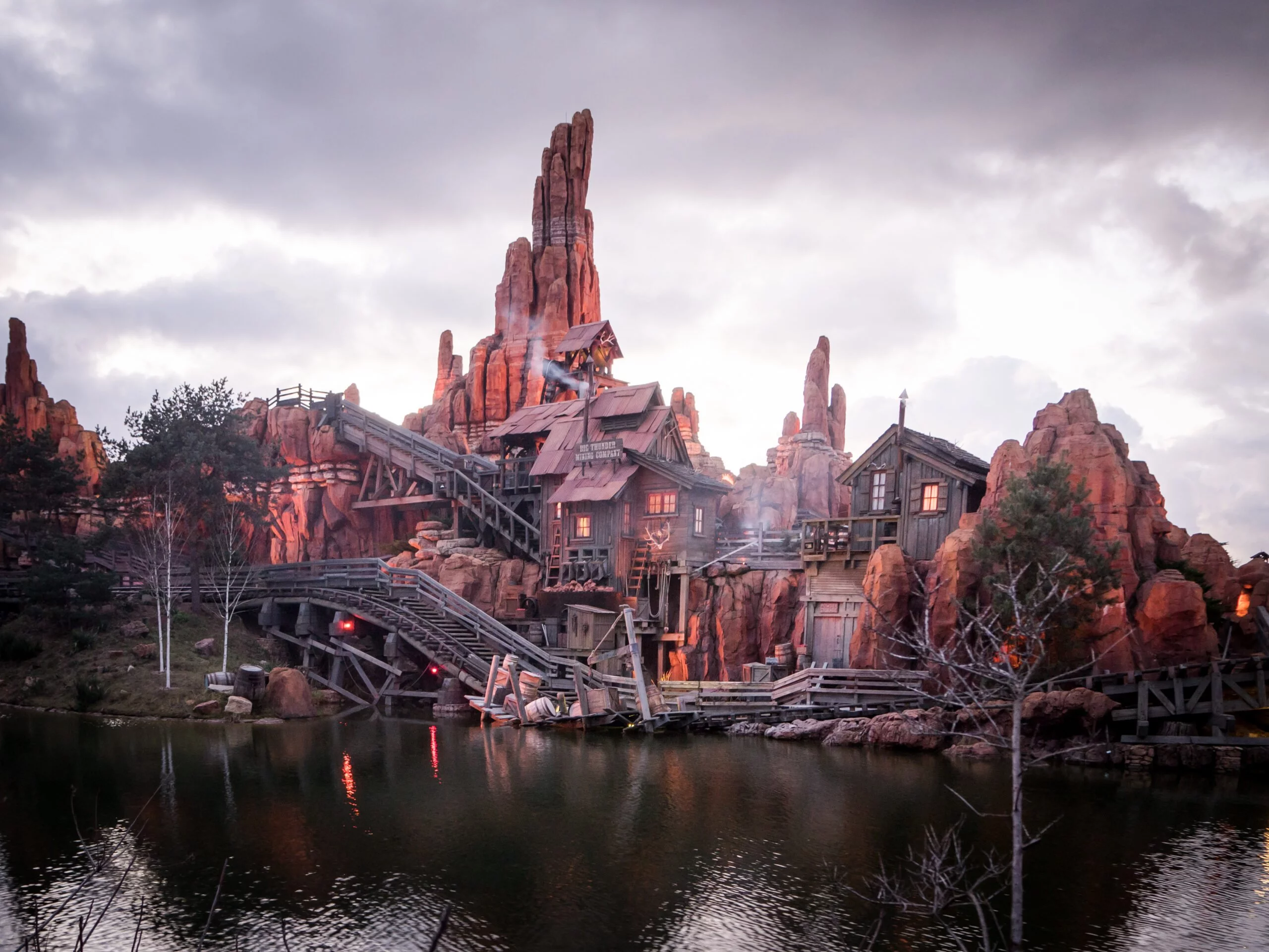 Une vue panoramique d'un paysage montagneux et accidenté avec un village en bois et des ponts au crépuscule, se reflétant dans un lac tranquille en contrebas, rappelant Disneyland Paris.