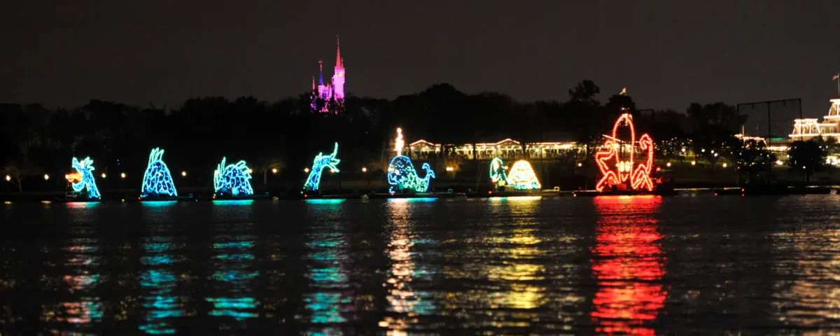 Une exposition nocturne de divers flotteurs thématiques illuminés sur l'eau, reflétant des couleurs vibrantes à la surface avec la silhouette d'un château et des arbres en arrière-plan, devient un spectacle aquatique électrique.