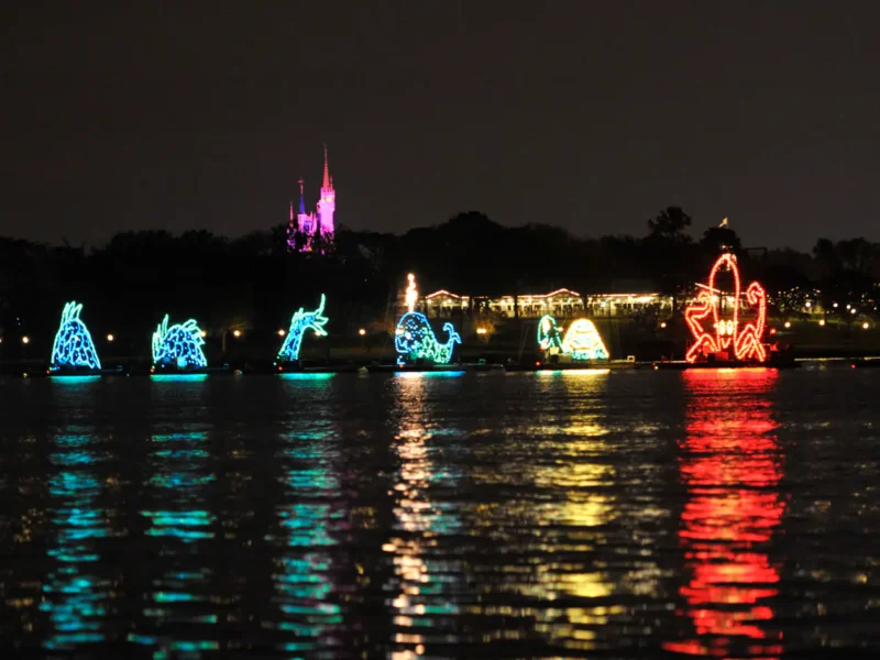 Une exposition nocturne de divers flotteurs thématiques illuminés sur l'eau, reflétant des couleurs vibrantes à la surface avec la silhouette d'un château et des arbres en arrière-plan, devient un spectacle aquatique électrique.