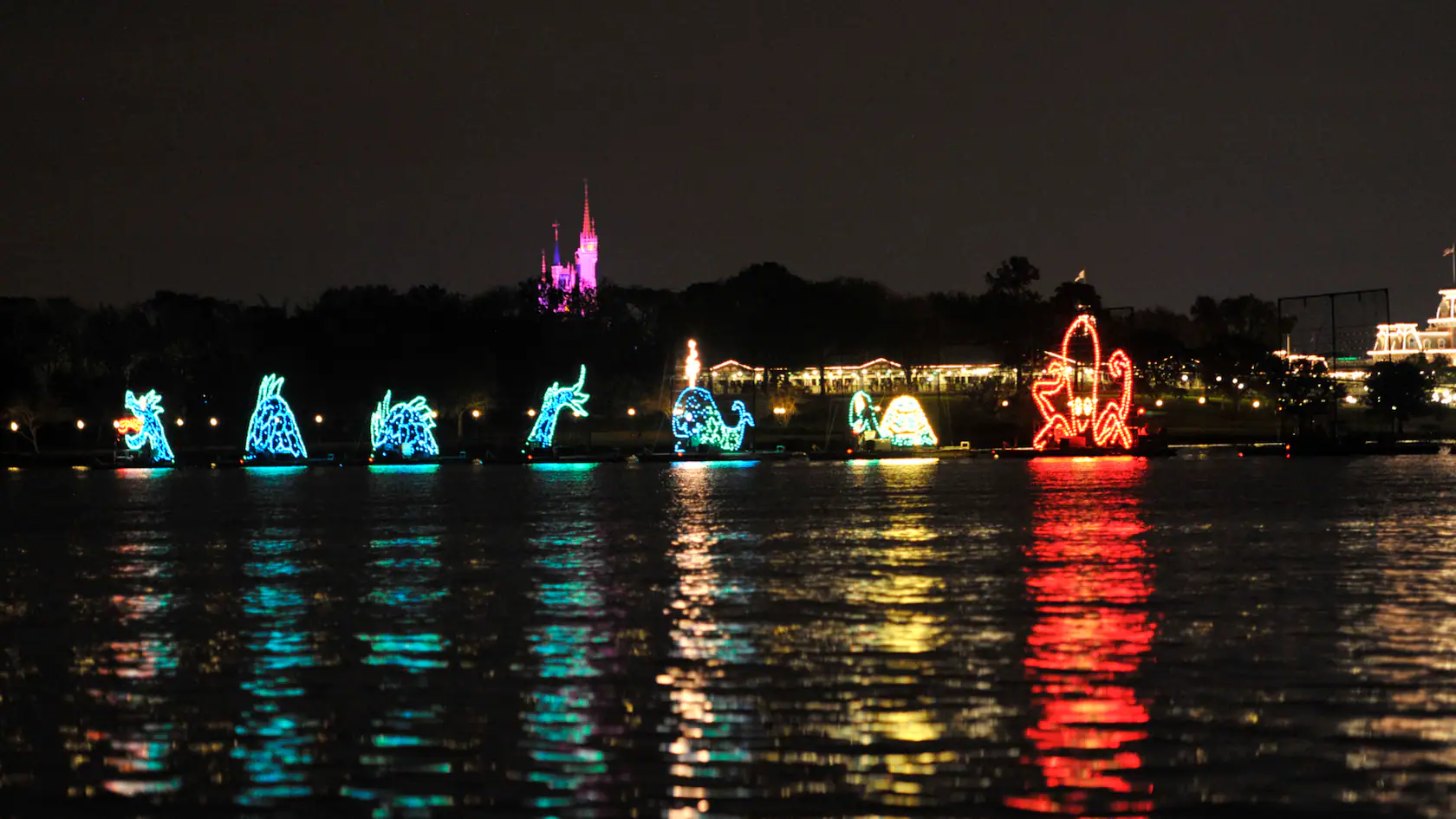 Une exposition nocturne de divers flotteurs thématiques illuminés sur l'eau, reflétant des couleurs vibrantes à la surface avec la silhouette d'un château et des arbres en arrière-plan, devient un spectacle aquatique électrique.