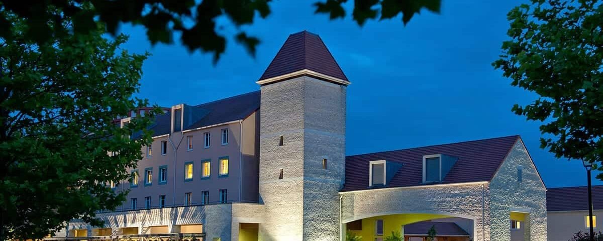 Un hôtel élégant pour les explorateurs au crépuscule, doté d'un design moderne avec une tour en pierre traditionnelle. Il est éclairé par un éclairage extérieur doux, vu à travers les feuilles d'arbres qui se détachent.