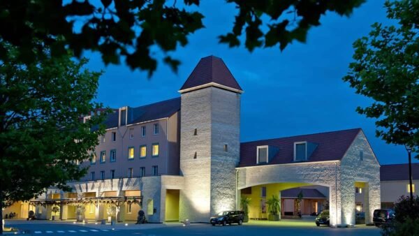 Un hôtel élégant pour les explorateurs au crépuscule, doté d'un design moderne avec une tour en pierre traditionnelle. Il est éclairé par un éclairage extérieur doux, vu à travers les feuilles d'arbres qui se détachent.