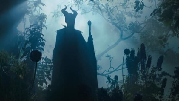 Une image sombre de Maléfique avec ses cornes emblématiques, vue de dos, regardant une forêt brumeuse mystique éclairée par un faisceau de lumière.