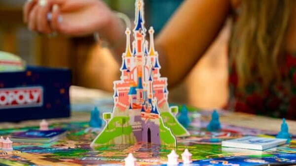 Un gros plan d'un jeu de société Monopoly coloré sur le thème de Disney avec un château en trois dimensions et des pièces de jeu miniatures, avec une femme jouant en arrière-plan.