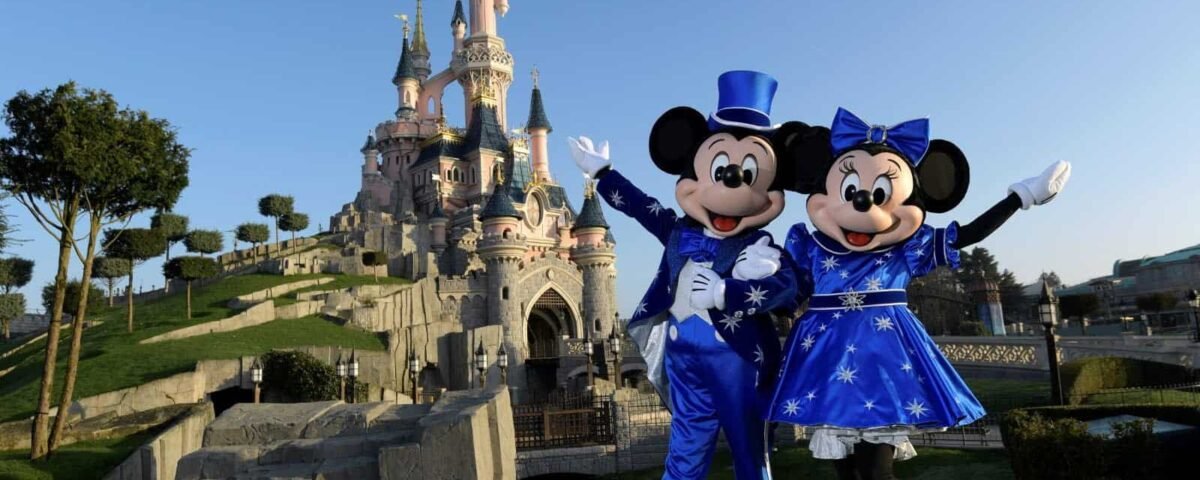 Mickey et Minnie Mouse en tenues bleues posant joyeusement devant le château de la Belle au bois dormant à Disneyland Paris par une journée ensoleillée.