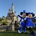 Mickey et Minnie Mouse en tenues bleues posant joyeusement devant le château de la Belle au bois dormant à Disneyland Paris par une journée ensoleillée.