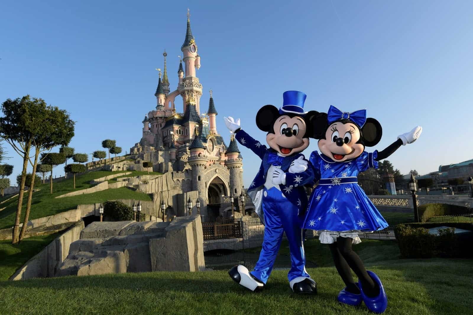 Mickey et Minnie Mouse en tenues bleues posant joyeusement devant le château de la Belle au bois dormant à Disneyland Paris par une journée ensoleillée.
