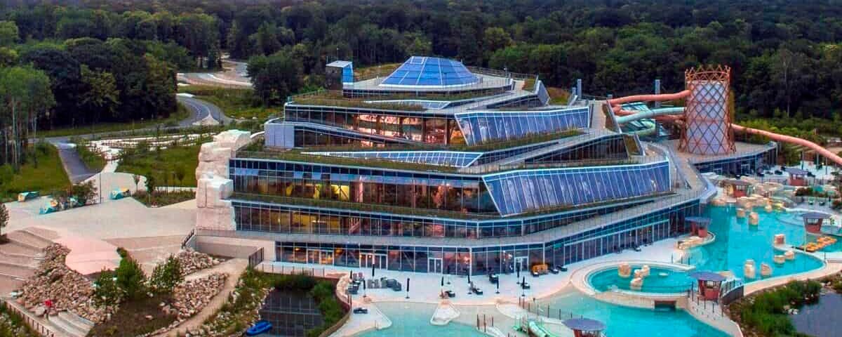 Vue aérienne d'un grand complexe aquatique moderne avec piscines intérieure et extérieure dans un village, entouré d'arbres et un coucher de soleil coloré en arrière-plan.