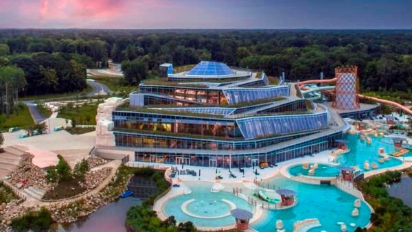 Vue aérienne d'un grand complexe aquatique moderne avec piscines intérieure et extérieure dans un village, entouré d'arbres et un coucher de soleil coloré en arrière-plan.