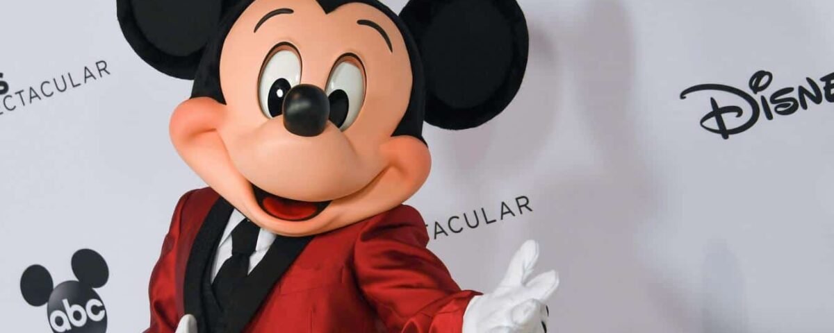 Mickey Mouse, vêtu d'un blazer rouge, pose joyeusement en agitant une main, sur un fond blanc orné des logos Disney et ABC.