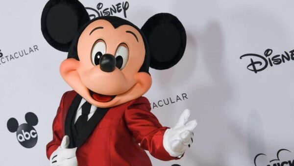 Mickey Mouse, vêtu d'un blazer rouge, pose joyeusement en agitant une main, sur un fond blanc orné des logos Disney et ABC.